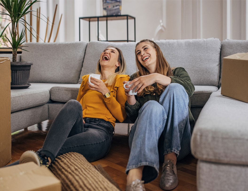 Two female friends sitting on living room floor surrounded by boxes.