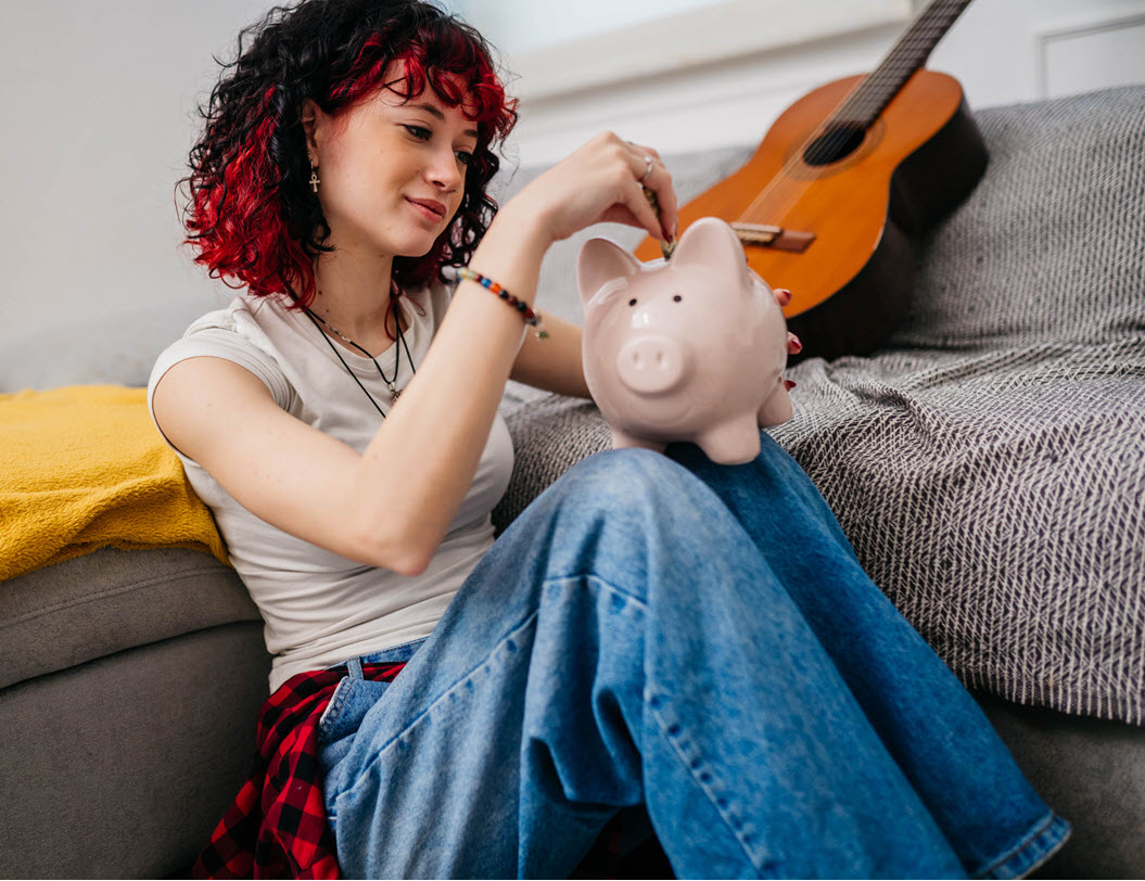 Teenage girl putting dollars in a piggy bank, guitar in the background on the couch